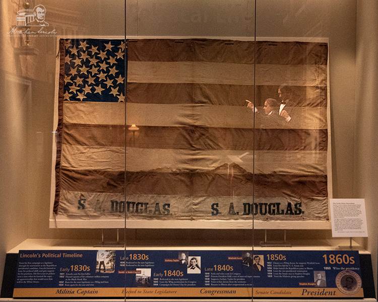 The banner in its display case, where a reflection from an exhibit showing the Lincoln-Douglas debates can be seen.